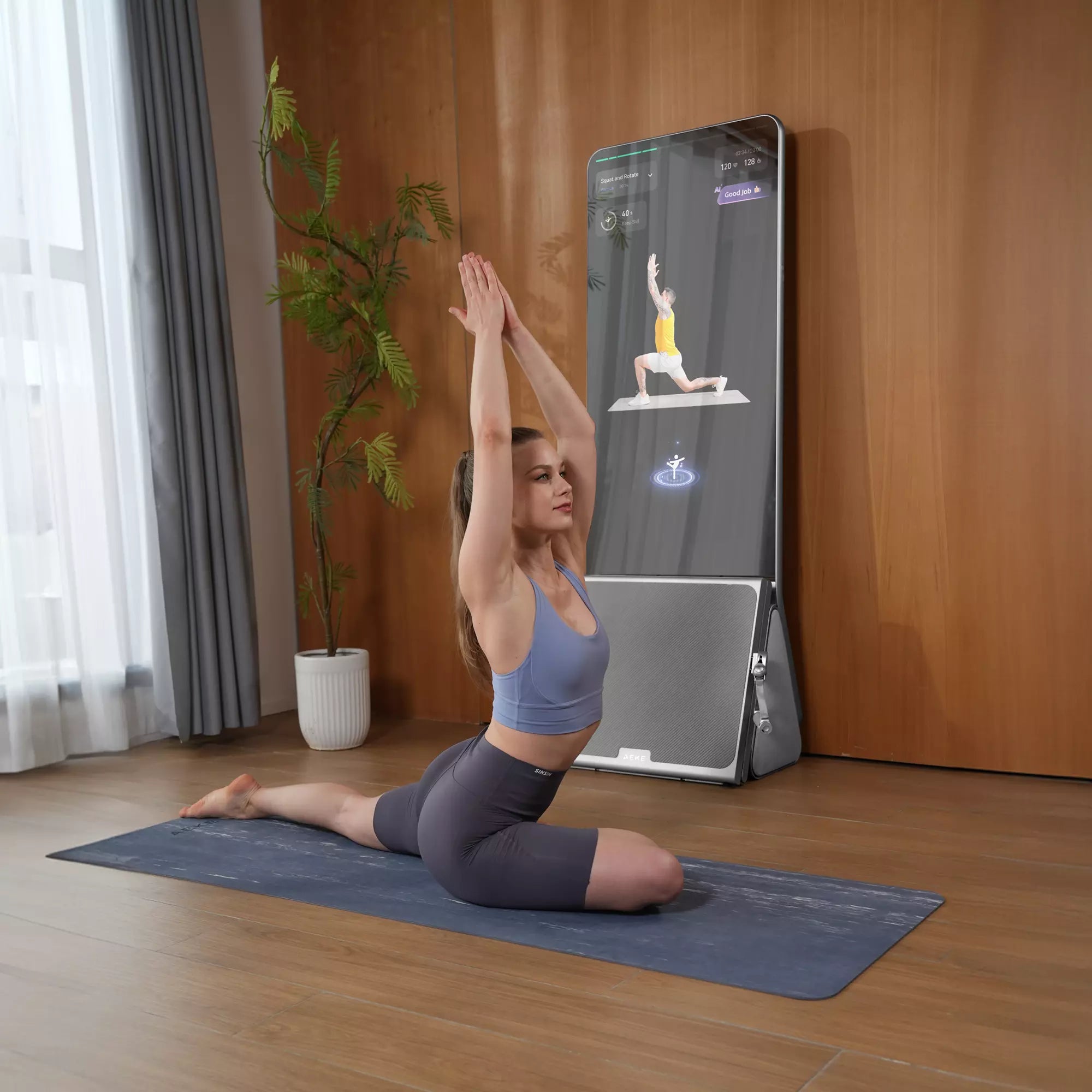 A woman follows a trainer on a tall fitness display while holding a stretch at home.