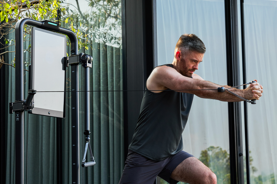 Man performing a standing cable chest press on a digital resistance training machine beside large windows