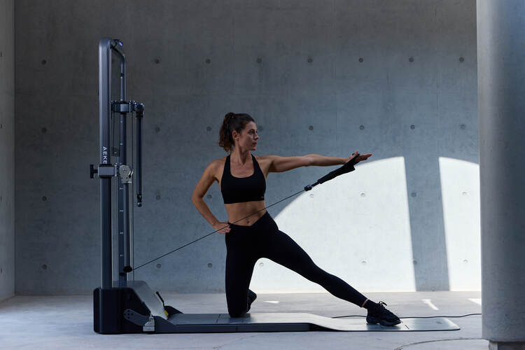 Woman standing beside a cable machine with one arm extended while holding a handle.